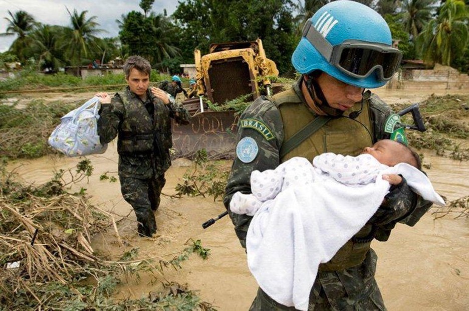 The image depicts two people wearing military camouflage uniforms in a flood-affected area. The person on the right is holding an infant wrapped in a white blanket with purple dots, and is wearing a blue helmet and goggles. The person on the left is carrying a bag of belongings over their shoulder. In the background, there is muddy floodwater with branches and debris scattered throughout. A bulldozer is visible behind them, and several palm trees and other trees are seen in the distance.